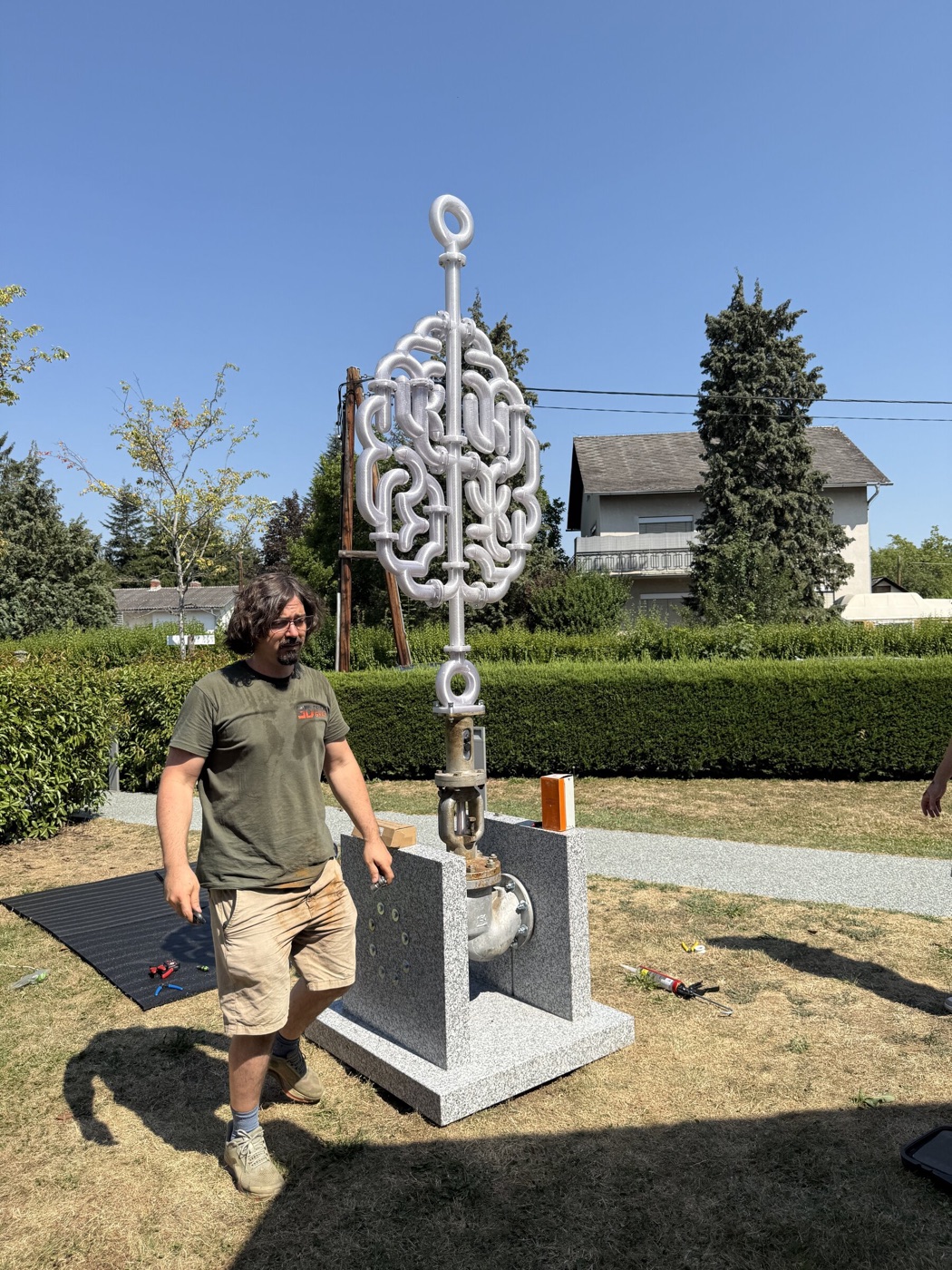 Die aufgestellte Skulptur auf ihrem Granitsockel vor Hecke und Himmel, Simon von 3D Druck Graz geht durchs Bild